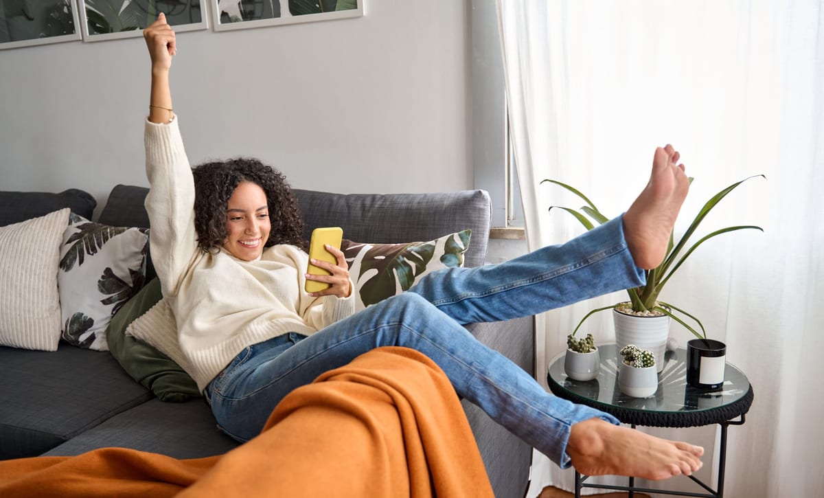 Happy excited young latin woman relaxing on couch using phone winning online.